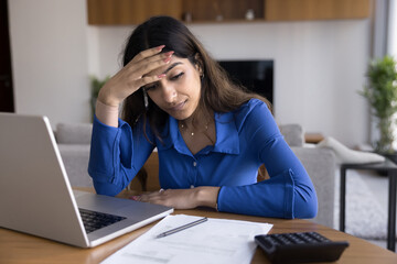 Worried sad young Indian homeowner woman thinking on money problems sitting at home workplace table with paper bills, invoices, calculator, laptop, touching head, looking at documents