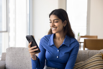Cheerful young adult Indian girl enjoying online communication on telephone at home, texting message on online chat, reading content on Internet, smiling, laughing, sitting on couch