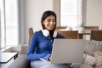 Happy young Indian freelance worker woman sitting on home couch with computer, using headphones, typing on laptop, chatting online, smiling, laughing. Student girl studying on Internet