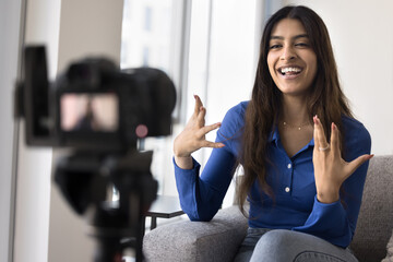 Cheerful Indian online influencer girl speaking at slr reflex camera on holder at home, filming video content for blog, vlog, social media, smiling, laughing, moving hands, giving training