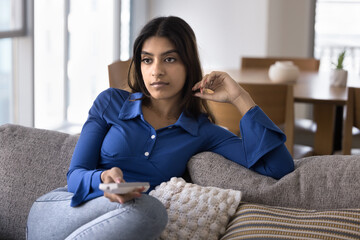 Serious attractive young 20s Indian girl watching movie, news broadcast, holding TV remote control device, browsing channels, feeling board, enjoying leisure, relaxing on home couch