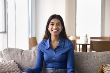 Cheerful beautiful young 20s Indian woman sitting on comfortable couch in cozy apartment, looking at camera with toothy smile, posing for home portrait. Influencer video call head shot