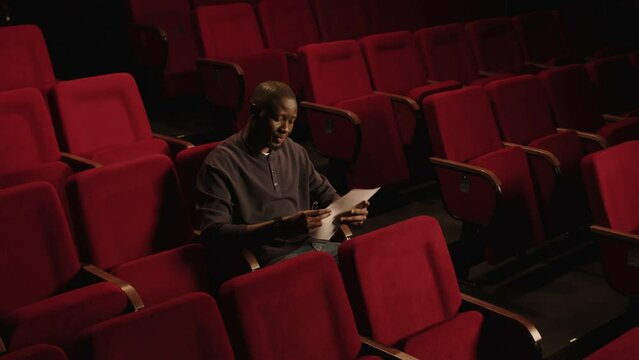 Pan shot of young Black male theatre actor holding sheets of script sitting on red seat and rehearsing for auditions