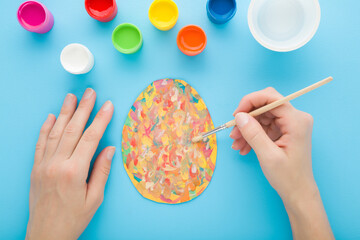 Young adult woman hand holding paintbrush and painting colorful egg on paper with watercolor. Pastel light blue table background. Closeup. Making easter decoration. Point of view shot. Top down view. - 785292672