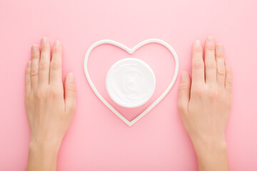 Young adult woman hands and white heart shape with cream jar on light pink table background. Pastel color. Care about clean and soft body skin. Closeup. Point of view shot. Top down view. - 785292695