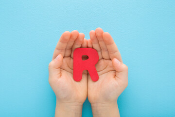 Baby boy hands holding red R letter on light blue table background. Pastel color. Time to learning. Closeup. Point of view shot. Top down view.
