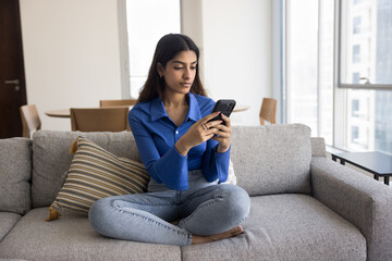 Serious young adult Indian smartphone user girl typing message on telephone, sitting on home couch, chatting, using smart service, app digital device for wireless Internet communication - 785292804