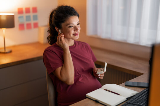 A smiling pregnant woman working from home and putting on her earphones to to listen to music