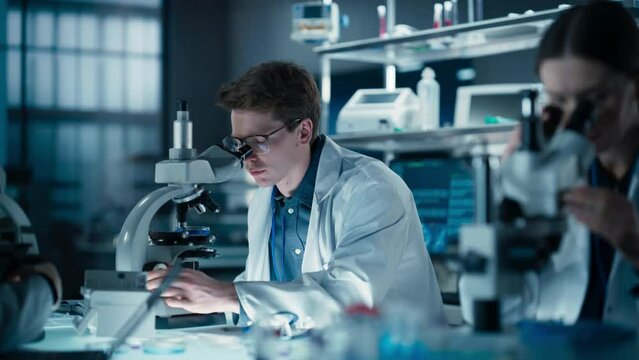 Diverse Male and Female Medical Research Scientists Work on a New Generation Medical Products in a Modern Laboratory. Focus on an Young Scientists Using Microscope, Analyzing Samples in a Petri Dish