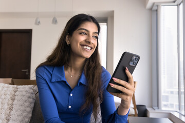 Happy dreamy young 20s Indian girl using smartphone on couch, holding gadget, looking away, smiling, thinking on Internet communication, smart home technology, laughing - 785295826