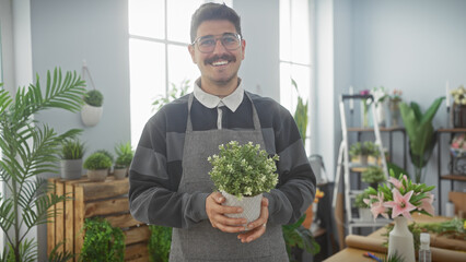 Smiling hispanic man with glasses and apron holding potted plant in flower shop with green plants...