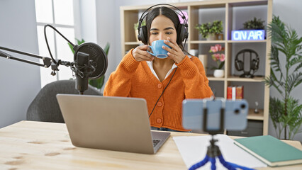 A young hispanic woman sips coffee in a radio studio setup with a microphone, laptop, and 'on air'...