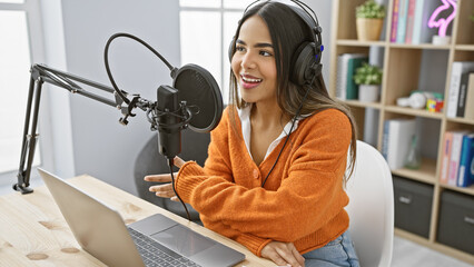 A smiling young hispanic woman records a podcast in a modern studio with a laptop and microphone.