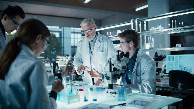 Young Team of Science Researchers Sitting Behind a Table, Using Microscopes for Medical Pharmaceutical Research and Treatment Development. Senior Scientist Supervising the Project