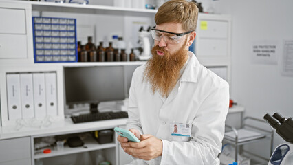 Handsome young redhead man, a serious scientist engrossed in his smartphone, indoors at a bustling...