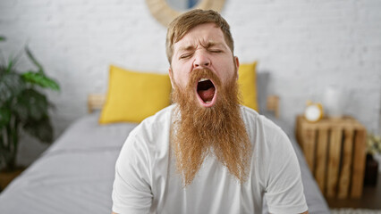 Exhausted young redhead man yawning hard, seated in bedroom, trying to wake up and start the morning