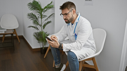 Handsome young hispanic man, a focused doctor, texting on his smartphone while sitting in a clinic...