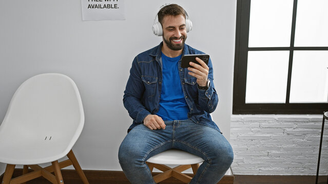 Handsome young hispanic man confidently immersed in vivid audio-visual world, sitting in waiting room chair, chilling, smiling as he's watching video and listening to music on smartphone.