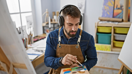 Young hispanic male artist, seriously focused at the art studio, drawing a portrait, while listening to music amidst brushes, canvas, and paint - obrazy, fototapety, plakaty