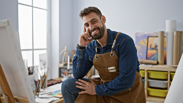 Portrait of a smiling young hispanic man, confidently sitting amid his art studio, brimming with paintings
