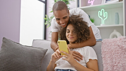 Beautiful couple confidently enjoying a fun-texting session, happily sitting on their home sofa,...