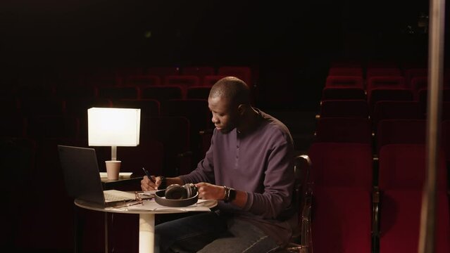 Medium shot of young African American male standup comedian sitting beside stage in amateur theater with retro red seats and writing monologue