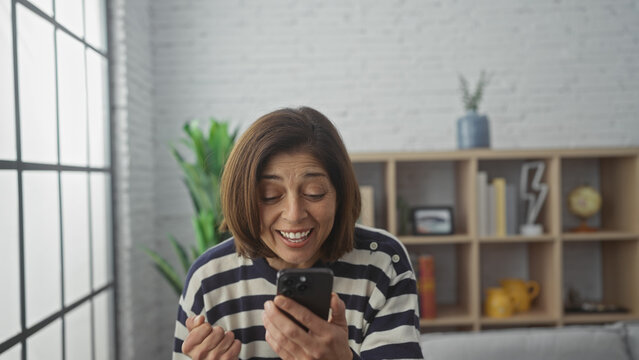 A mature hispanic woman laughs while using a smartphone in a cozy living room setting.