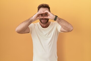 Fototapeta na wymiar Middle age man with beard standing over yellow background doing heart shape with hand and fingers smiling looking through sign