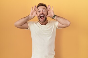 Middle age man with beard standing over yellow background smiling cheerful playing peek a boo with...