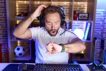 Middle age man with beard playing video games wearing headphones looking at the watch time worried,...