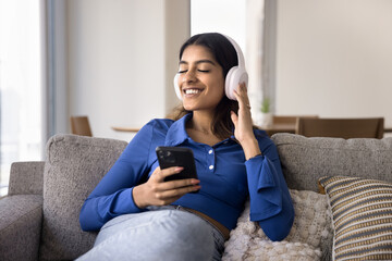 Happy peaceful young Indian student girl listening to music from wireless headphones with closed eyes, enjoying favorite relaxing ambient tunes, holding smartphone, resting on home couch