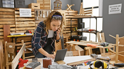 Portrait of a beautiful young hispanic woman carpenter, engrossed in online business on her touchpad while casually talking on smartphone amidst the rustic charm of her carpentry workshop - obrazy, fototapety, plakaty
