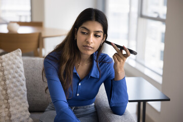Serious focused young adult Indian girl listening to voice audio message on telephone, holding smartphone dynamic at ear, thinking, looking away in deep thoughts