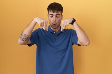 Young hispanic man standing over yellow background pointing down with fingers showing...