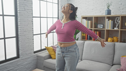 Joyful young hispanic woman dancing in a cozy living room with modern decor. - obrazy, fototapety, plakaty