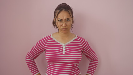 A young hispanic woman with glasses posing against a pink wall, exhibiting a confident stance.