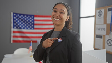 A smiling hispanic woman points to her 'i voted' sticker in a college voting center with an american flag backdrop