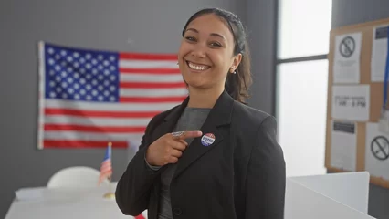 Poster A smiling hispanic woman points to her 'i voted' sticker in a college voting center with an american flag backdrop © Krakenimages.com