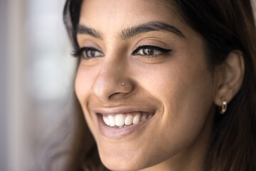 Cheerful beautiful young Indian woman face close up. Positive beauty care model girl with smooth perfect facial skin, dental patient with toothy smile looking at away, posing for macro portrait