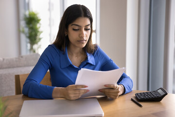 Serious focused young Indian woman reading legal financial document, doing accounting paperwork at home workplace table with calculator and laptop, checking paper reports, bills