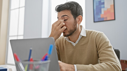 Stressed hispanic man in office rubbing eyes at desk with laptop
