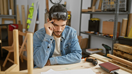 A thoughtful hispanic man with a beard wearing safety glasses reflects in a cluttered carpentry...