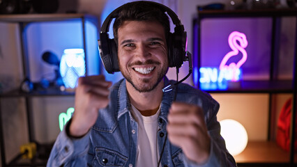 Happy hispanic man wearing headphones in a neon-lit gaming room at night, embodying modern...