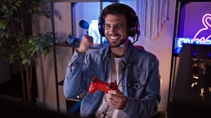 A jubilant young hispanic man with a beard celebrates a gaming victory in a dark room at night.