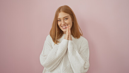A cheerful young caucasian woman with red hair in a cozy white sweater posing against a soft pink...
