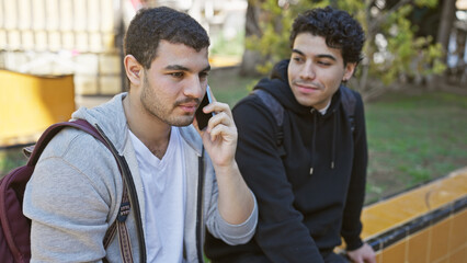 Two hispanic men in a park, one talking on a phone and the other looking on, depict friends in a...