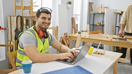 Two men in a carpentry workshop, one smiling at the camera while using a laptop and the other...