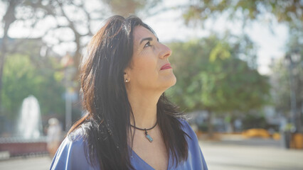 A middle-aged hispanic woman gazes contemplatively outdoor in a sunny, green park.