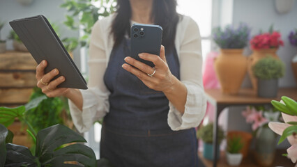 A woman uses a tablet and smartphone in a flower shop surrounded by colorful plants and pottery,...