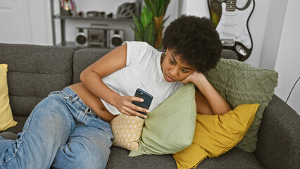 African american woman using smartphone on couch indoors with guitar in the background - obrazy, fototapety, plakaty
